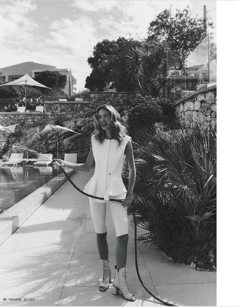 Black and white fashion shot of a woman in an elegant white outfit with a hose on a sunny stone patio beside a pool.