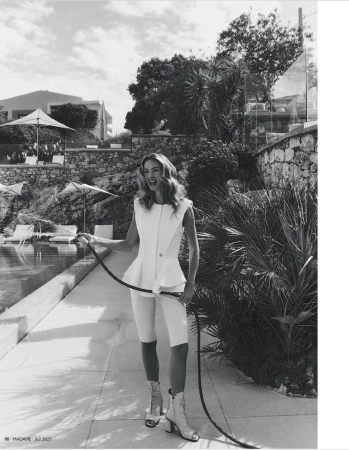 Black and white fashion shot of a woman in an elegant white outfit with a hose on a sunny stone patio beside a pool.