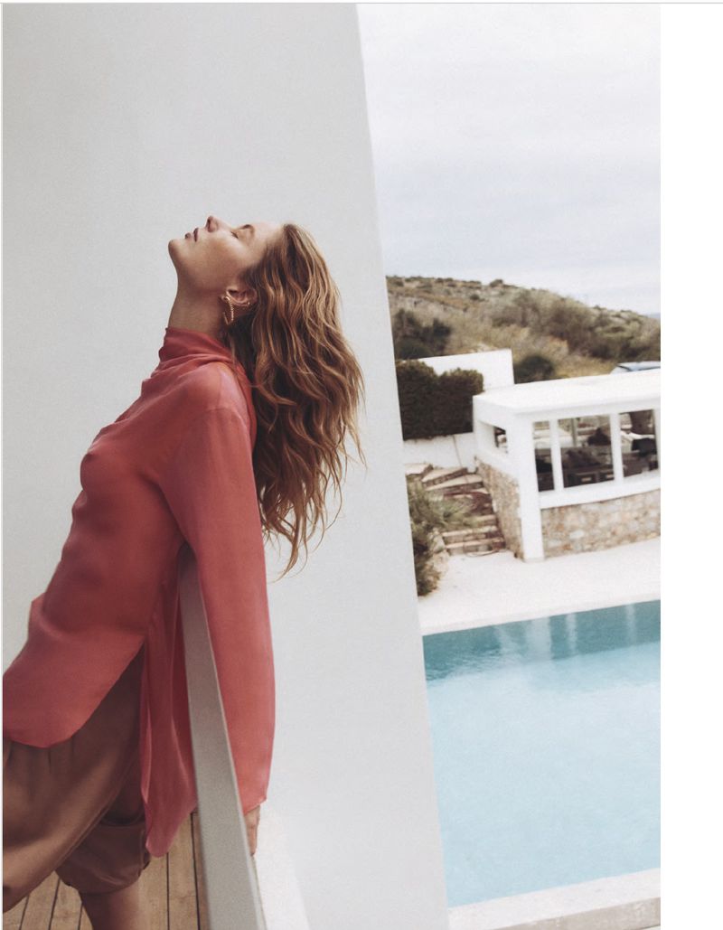 Outdoor portrait of a young adult woman in coral shirt leaning on a railing next to a turquoise pool with houses in the background.