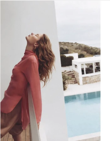 Outdoor portrait of a young adult woman in coral shirt leaning on a railing next to a turquoise pool with houses in the background.