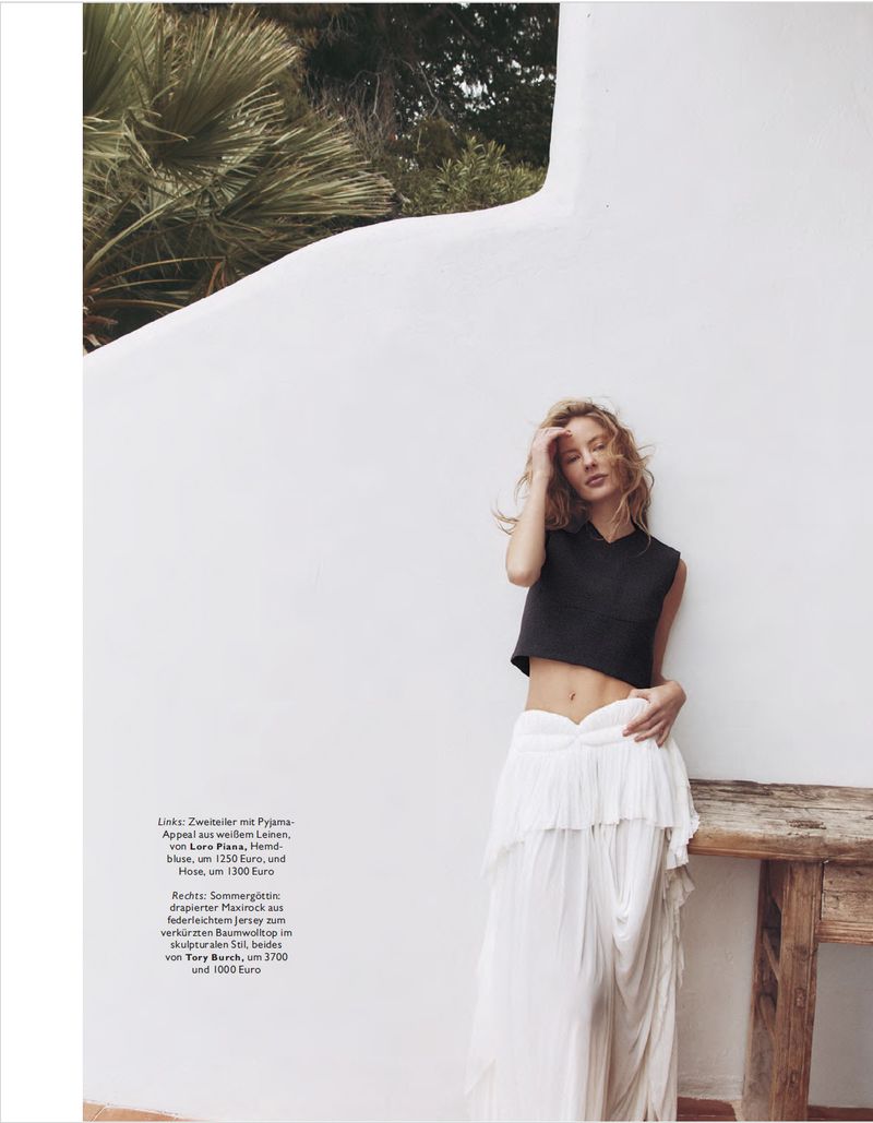 Fashion editorial shot of a blonde model in a black crop top and white maxi skirt posing against a textured white wall and greenery.