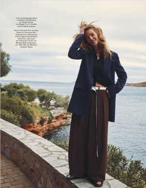 Model standing on stone wall by the sea wearing an oversized blue blazer and brown trousers with white belt