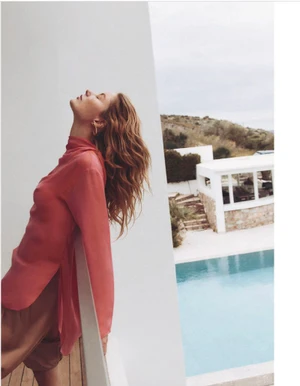 Model wearing a pink top leaning against a white balcony overlooking a swimming pool and coastal buildings