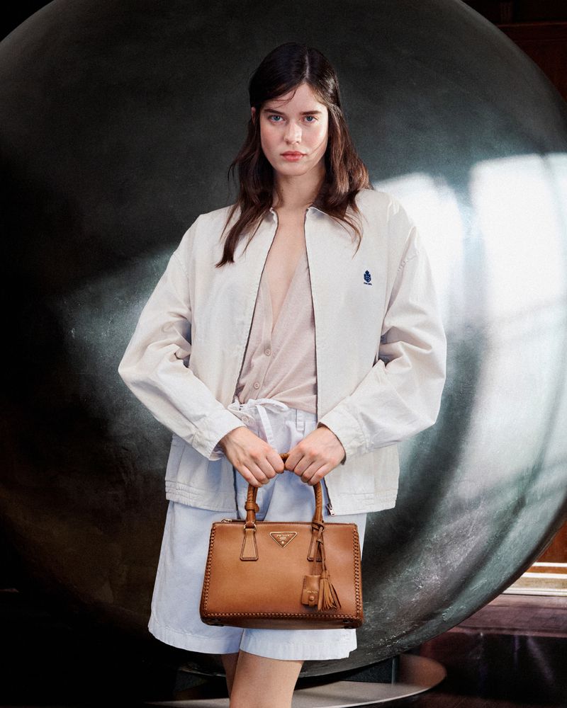 Fashion shot of a young adult woman with dark hair in a white jacket and light blue shorts, holding a tan Prada handbag.