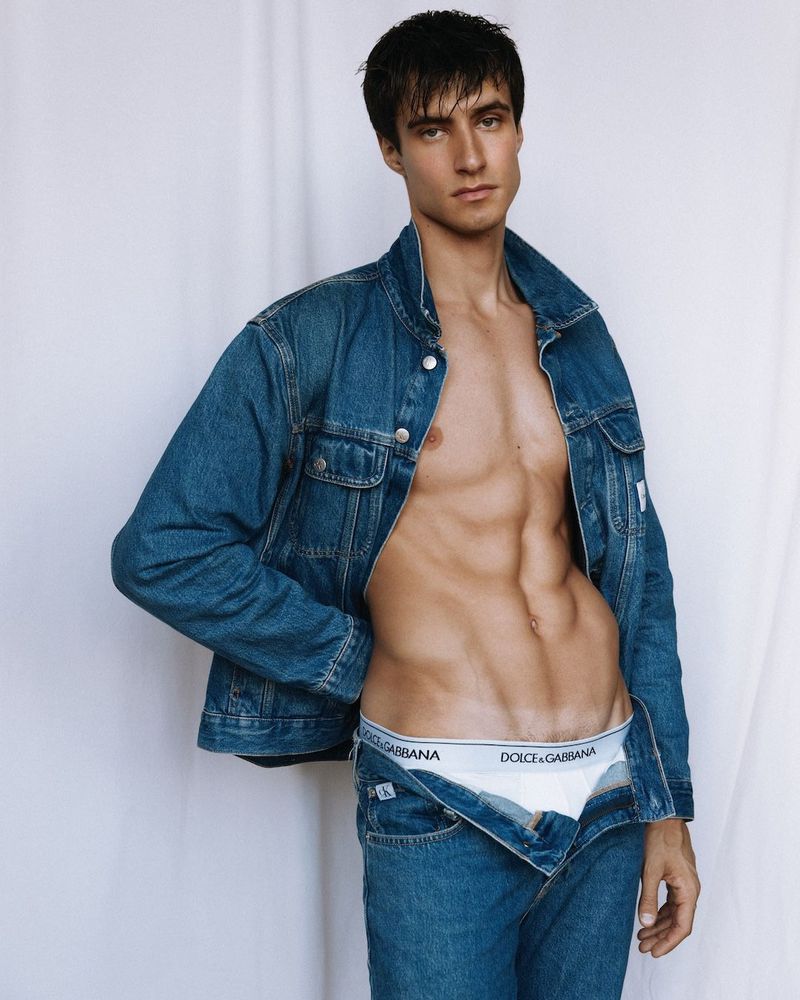 Man poses shirtless in denim jacket and jeans, showcasing muscular physique, with a neutral expression against a bright, clean backdrop.