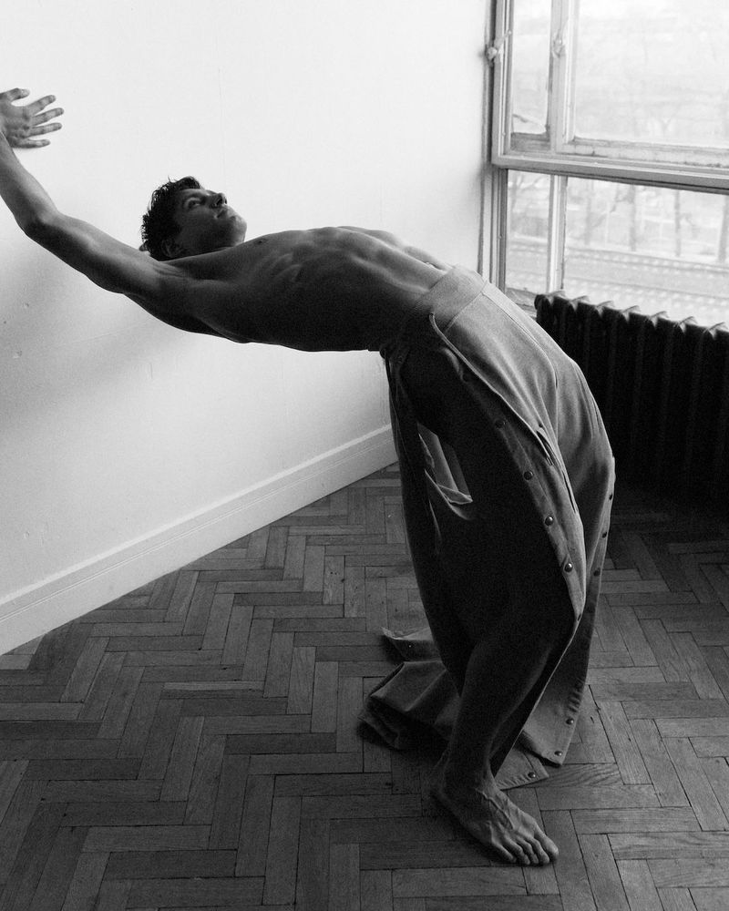 Black and white shot of a muscular man in pants bending backwards against a white wall near a window, showcasing his physique and form.