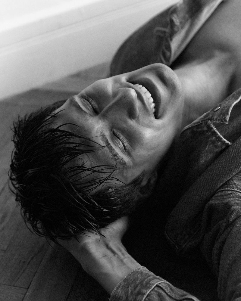 Black and white portrait of an adult laying down with a happy expression, wearing a denim shirt, captured with eye-level framing.