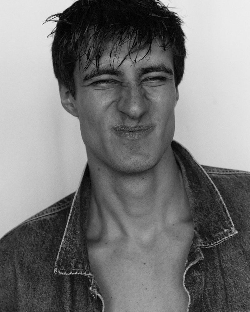 Monochrome portrait of a young adult man with a scrunched face and open denim jacket against a white wall backdrop, close-up shot.