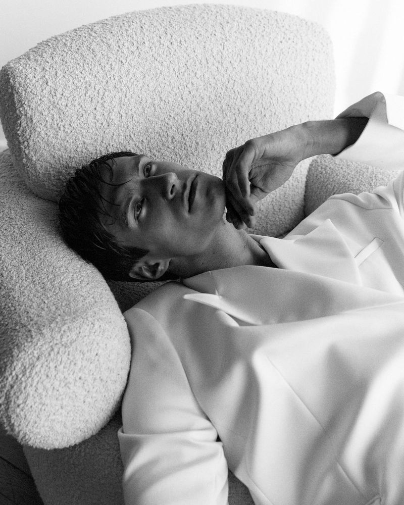 Black and white studio shot of an adult male lying on a white fluffy chair, wearing a white blazer and looking up.