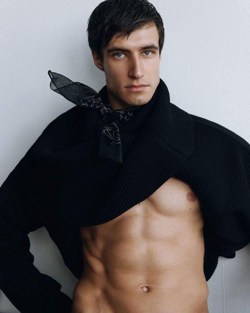 Portrait of a fair-skinned adult man with dark hair, wearing a black sweater and a black bandana, showcasing his muscular physique in a studio.