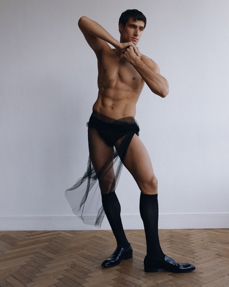 Shirtless adult male in artistic pose wearing black underwear, tulle skirt, socks, and loafers against a neutral backdrop and wood floor.