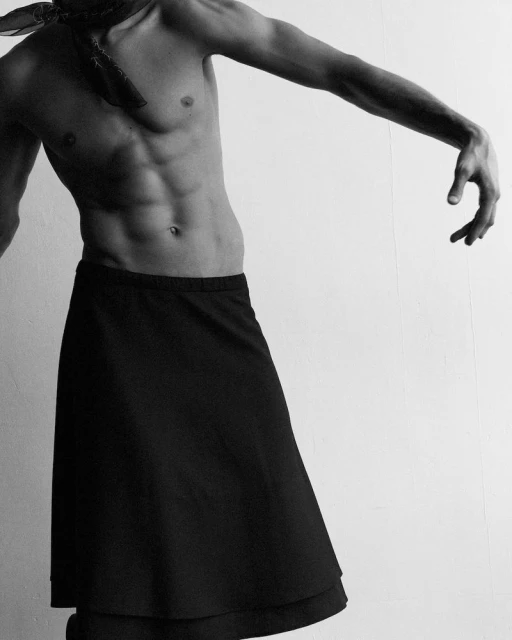 Monochrome studio shot of a shirtless adult male with a black skirt, flexing abs against a white backdrop.