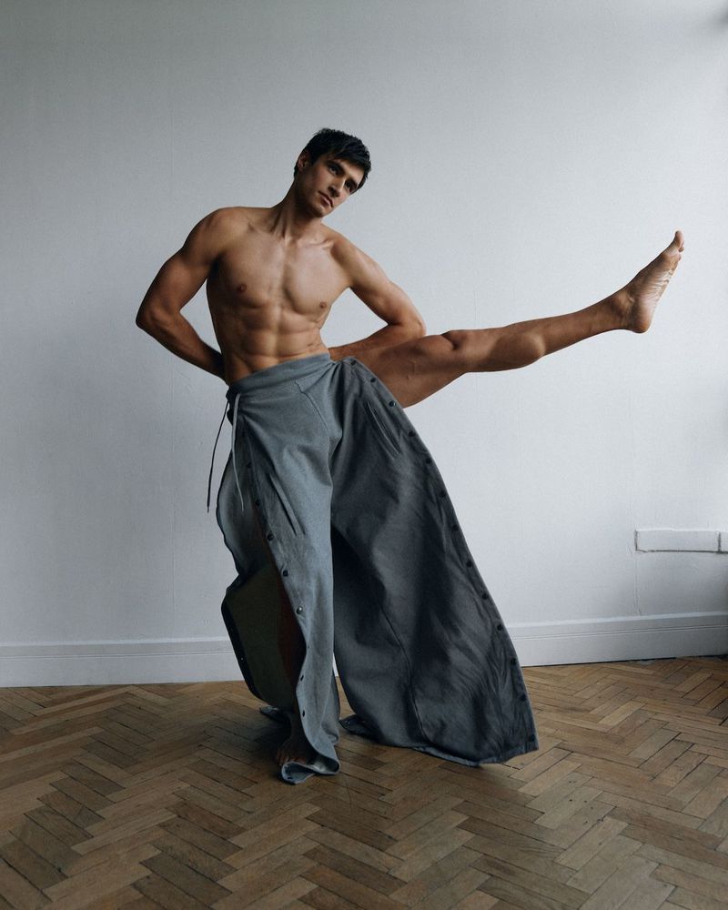 Shirtless adult male with dark hair and muscular build is striking a dynamic pose with legs extended against a plain white background.