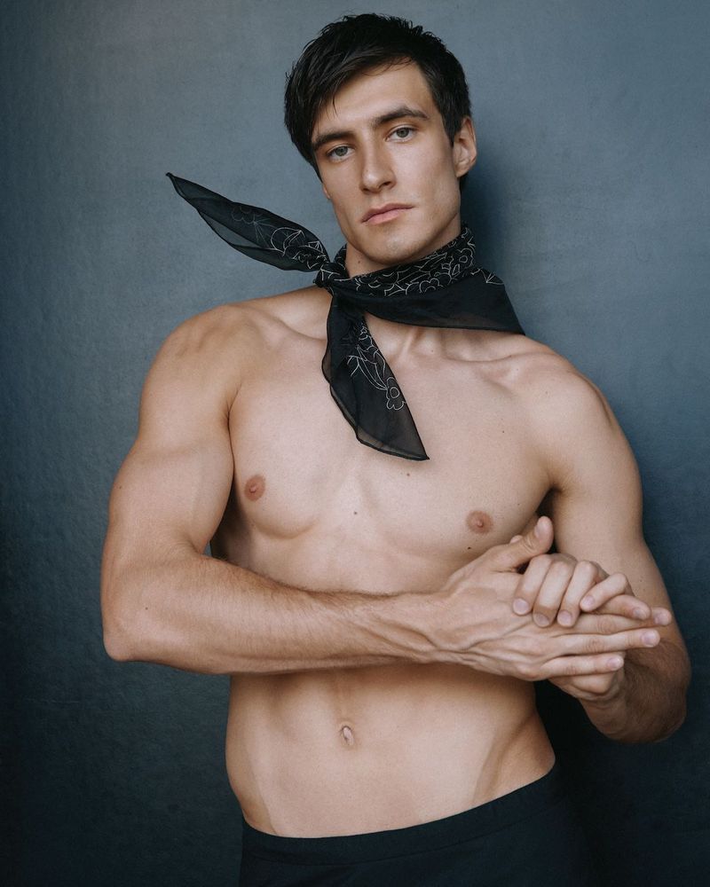 Shirtless man with muscular build posing in a studio with a black patterned bandana tied around his neck, standing against dark background.