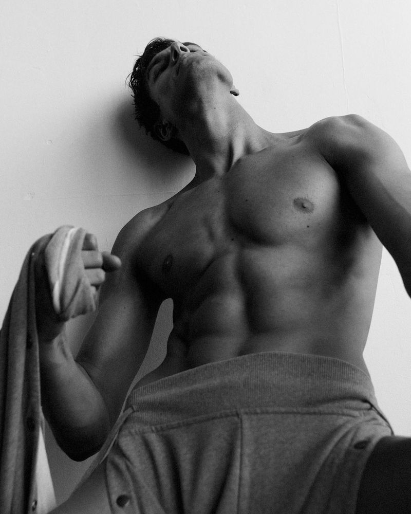 Low-angle black and white shot of muscular adult male torso with partial face, wearing gray sweatpants, holding the matching jacket.
