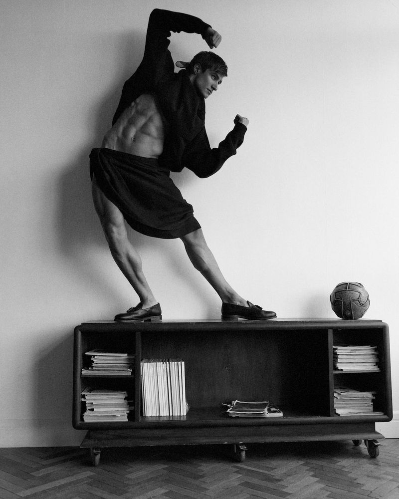 Black and white portrait of a muscular man in skirt and jacket standing on a mid-century modern console with books and a soccer ball.