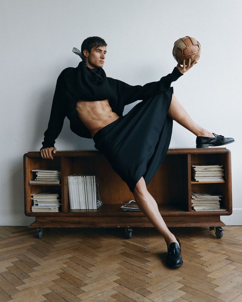 Attractive adult male balancing a vintage soccer ball on his foot while posing next to a mid-century modern console in a fashion editorial.
