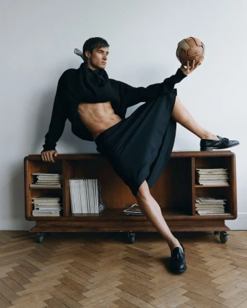 Attractive adult male balancing a vintage soccer ball on his foot while posing next to a mid-century modern console in a fashion editorial.