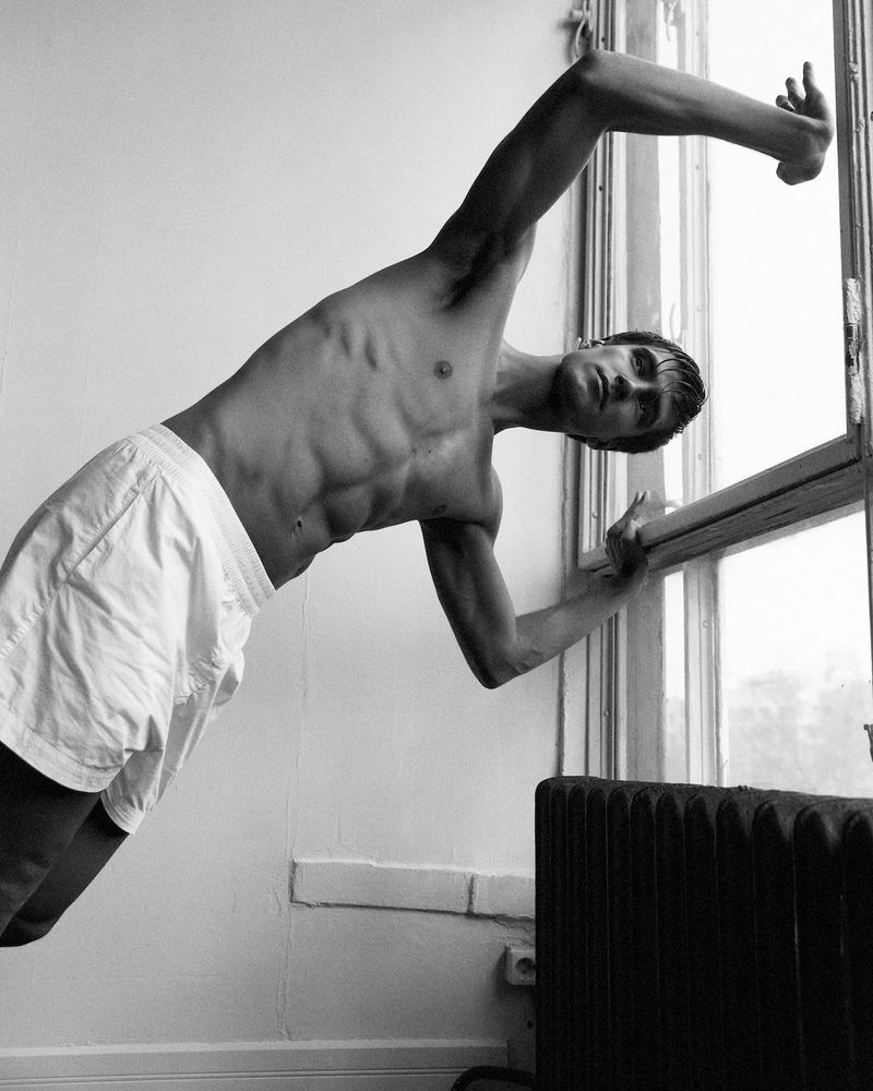 Black-and-white studio shot of a shirtless man in white shorts, leaning against a window with a radiator in the corner. 