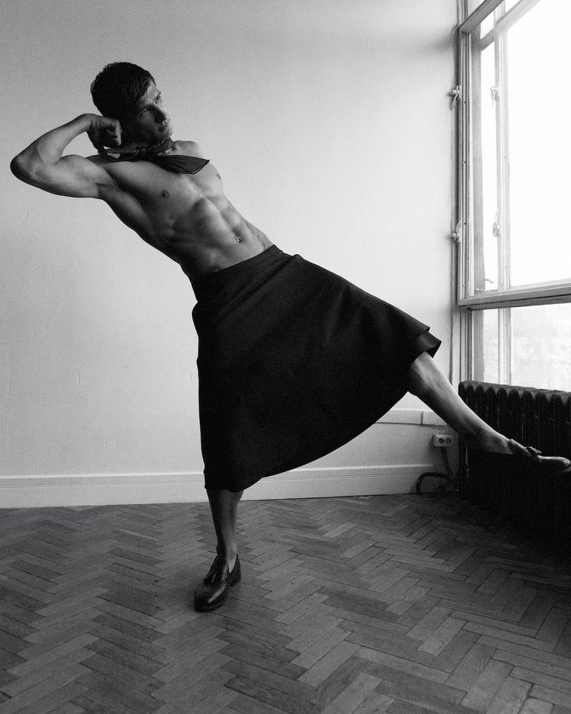 Black and white studio fashion shot of a muscular man in a skirt posing by a window with natural light on a herringbone floor.