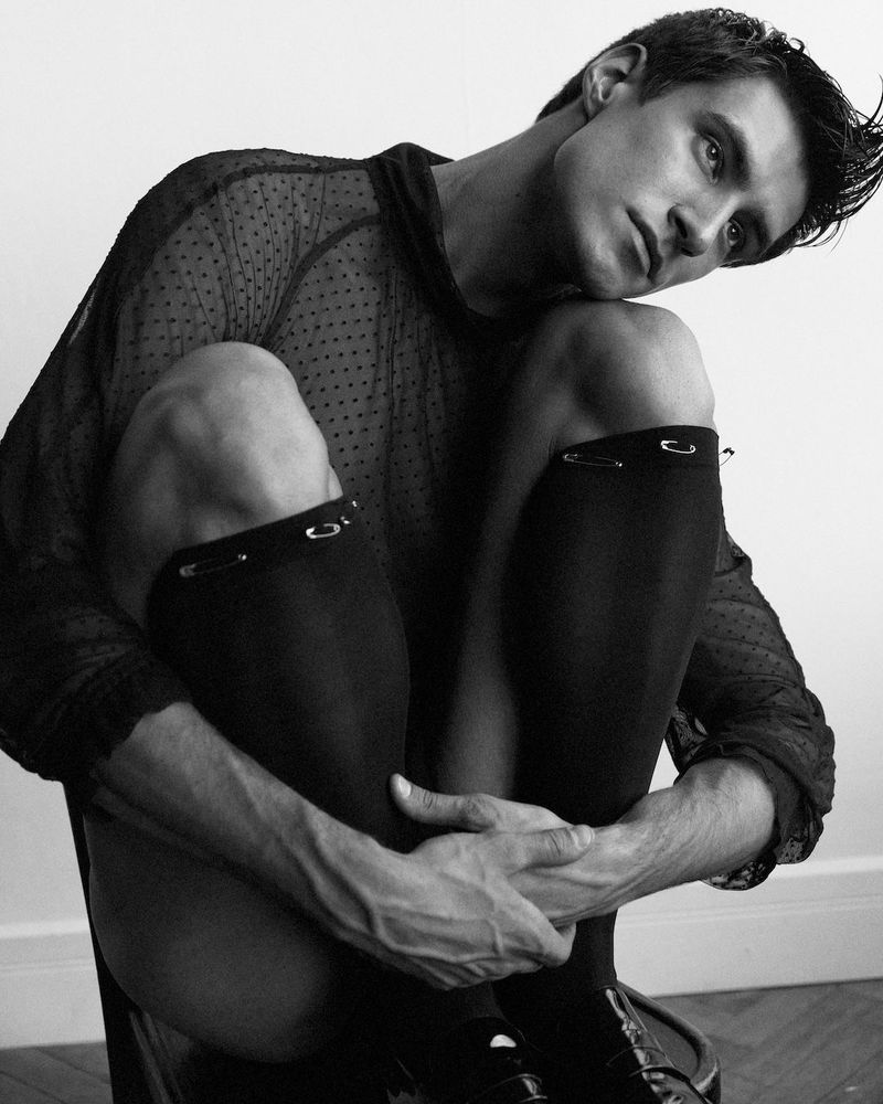 Black and white studio portrait of a young adult male model with dark hair, wearing a sheer black shirt, socks with safety pins, and shoes.