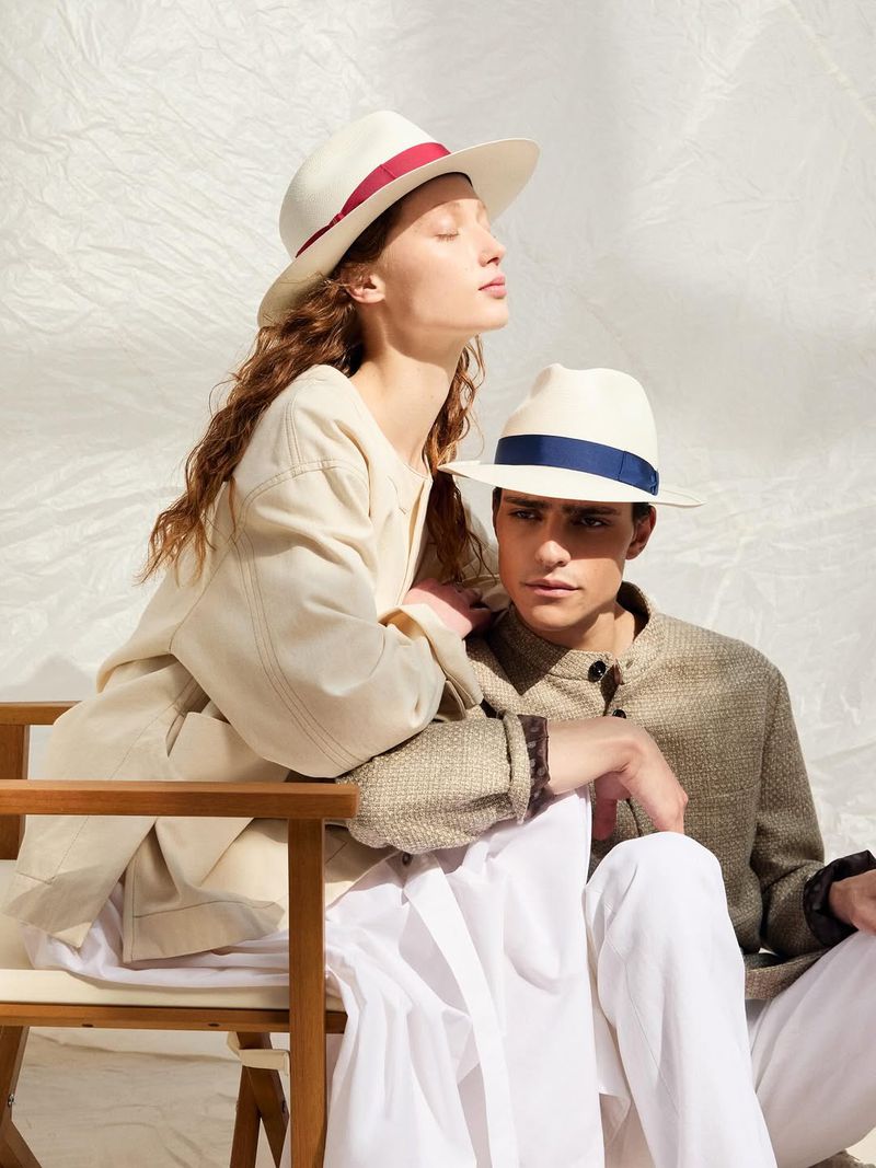 Fashion photo featuring a woman in a cream-colored hat and jacket and a man in a hat and brown jacket in a studio setting.