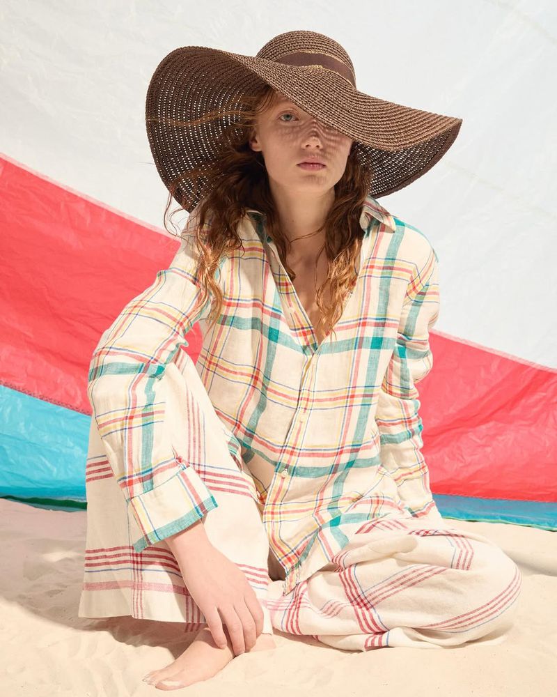 Model wearing a wide-brim brown straw hat and plaid shirt sitting on a sandy beach