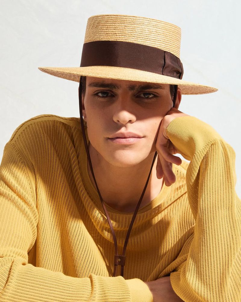 Man wearing a straw boater hat with a brown ribbon and a yellow ribbed sweater against a plain white wall