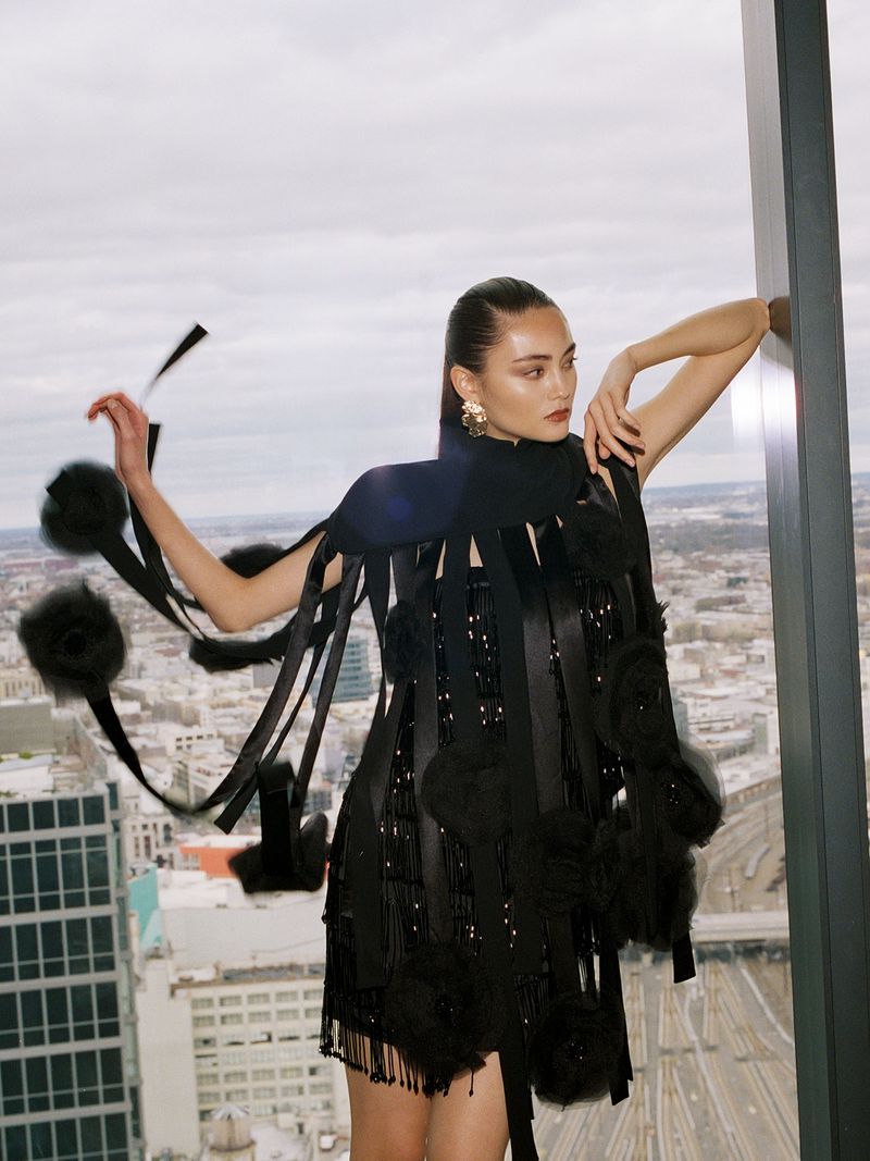 Fashion shot: Woman in avant-garde black dress with ribbons and pom poms poses near city view window. Editorial fashion concept.