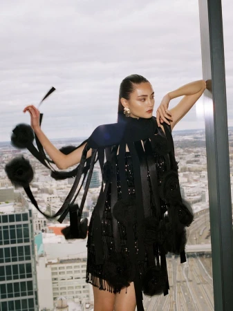 Fashion shot: Woman in avant-garde black dress with ribbons and pom poms poses near city view window. Editorial fashion concept.