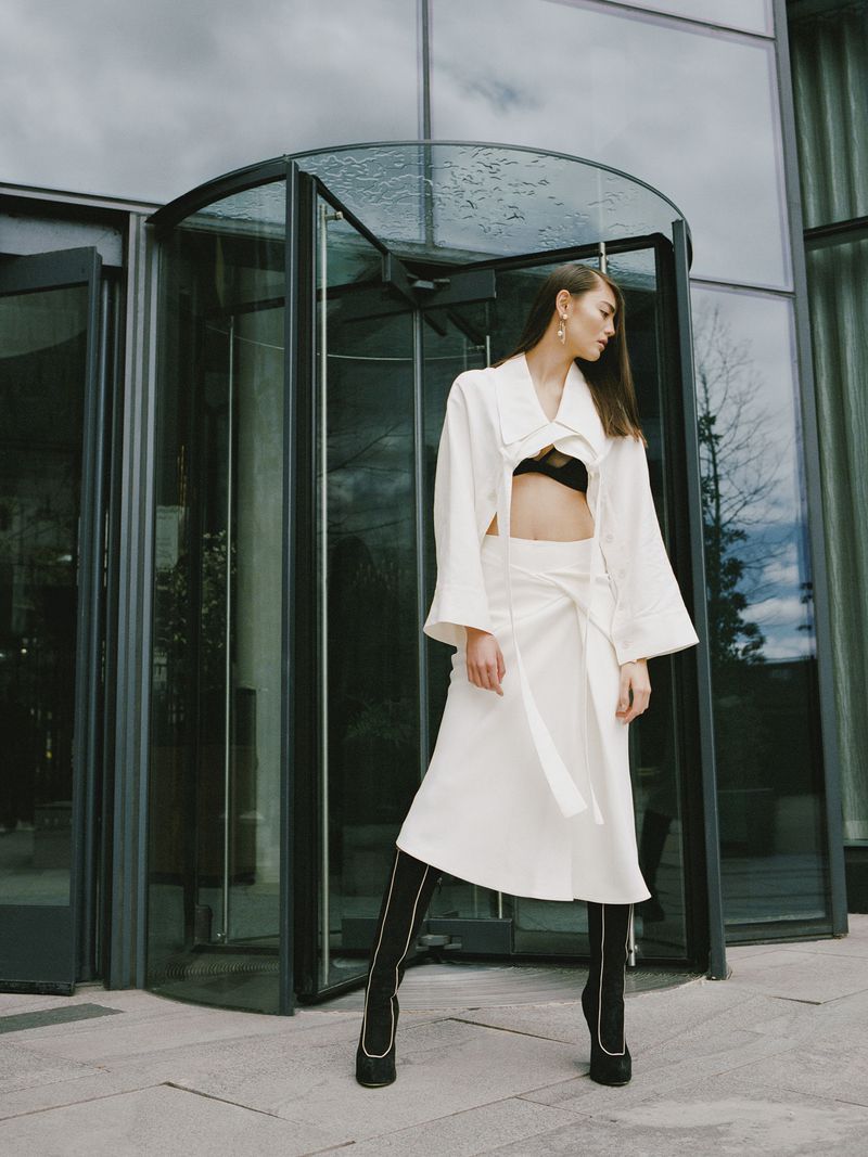 Fashionable woman in white skirt and jacket poses confidently near a modern glass building entrance, showcasing contemporary style and design.