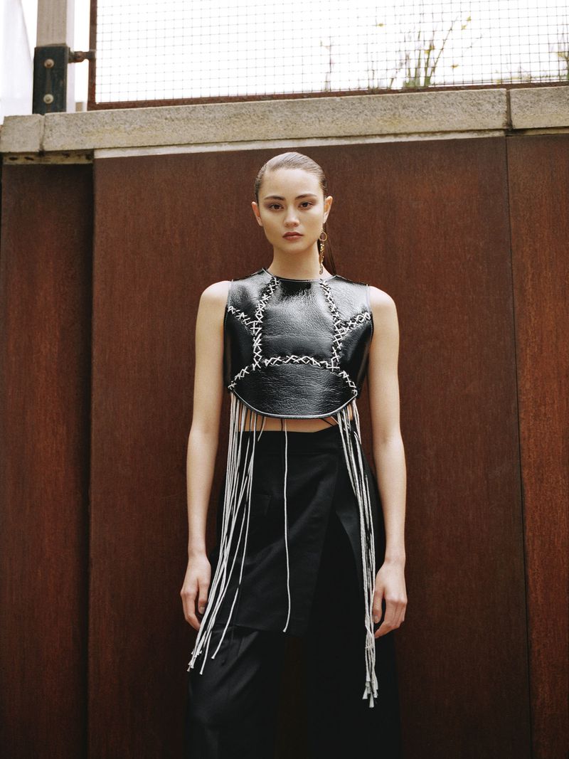 A young woman poses in a black leather top with fringe and a black skirt against a rusty metal background in a fashion-style portrait.