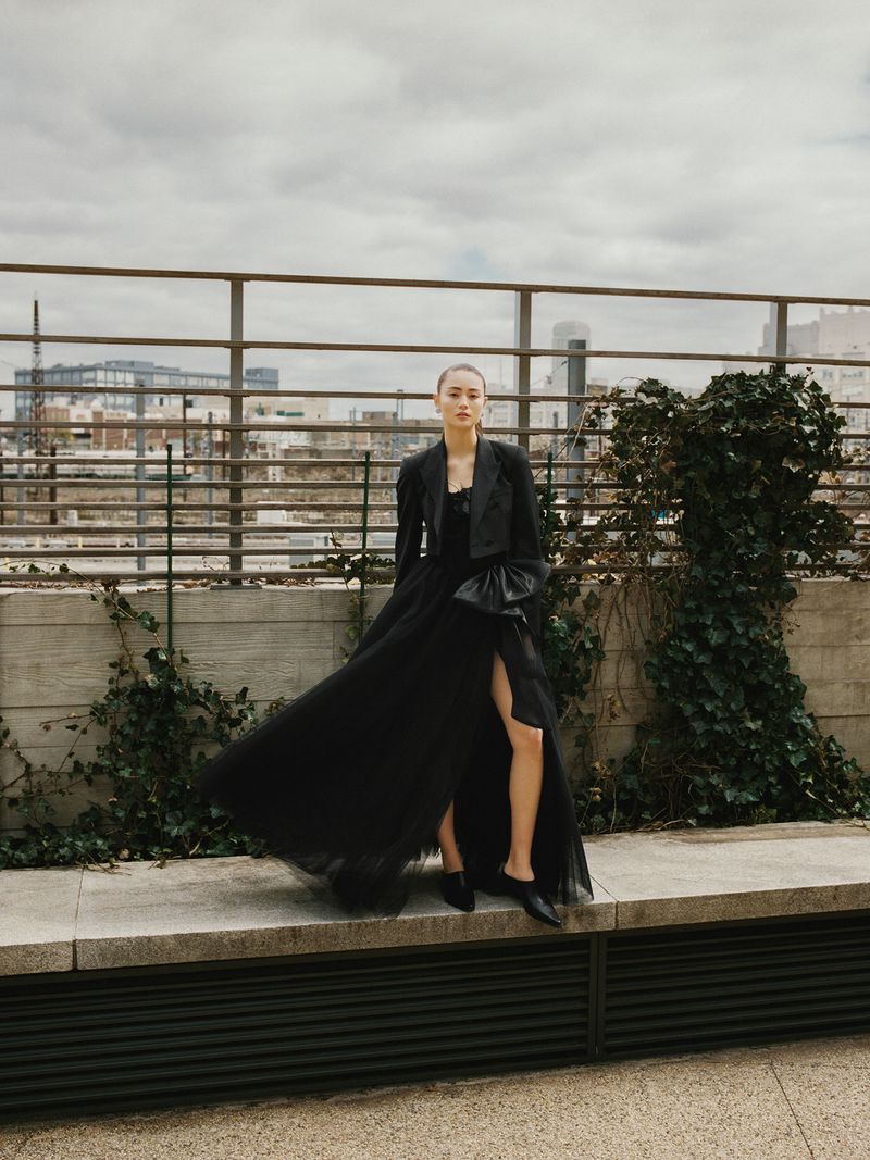 Fashionable woman in a black gown poses on a rooftop with city views and greenery, showcasing a stylish and elegant outdoor shot.