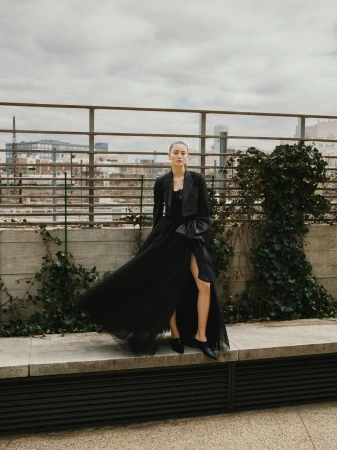 Fashionable woman in a black gown poses on a rooftop with city views and greenery, showcasing a stylish and elegant outdoor shot.