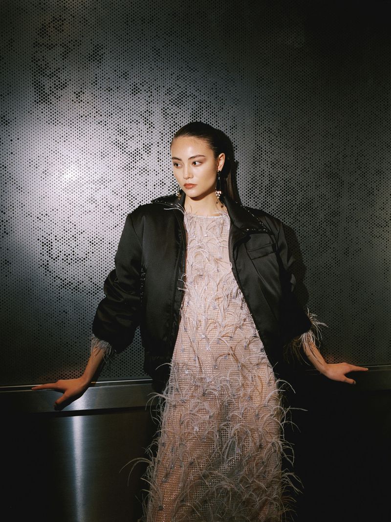 A stylish woman posing indoors with a black bomber jacket and feather dress, leaning against a metallic textured wall for a fashion shot.
