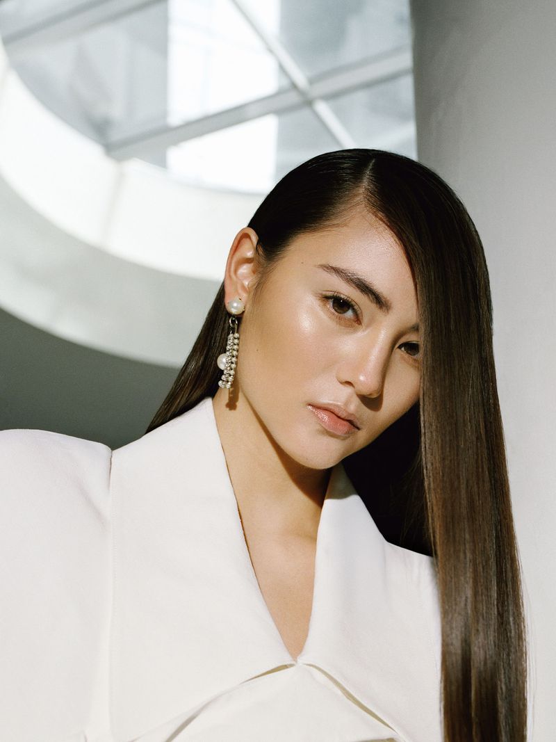 Close-up of an adult female with straight brown hair wearing an elegant white jacket with pearl earrings against a white background.