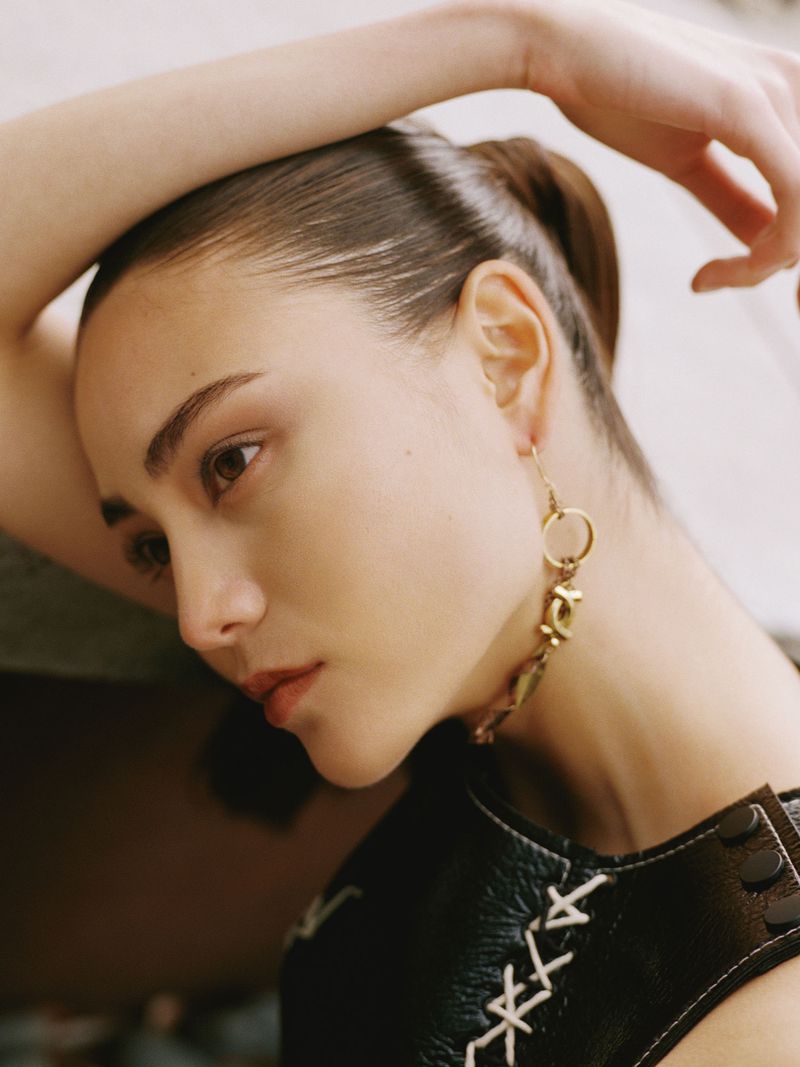 Close-up portrait of a young fair-skinned woman with dark hair, wearing statement earrings, and a black leather top with white stitching.