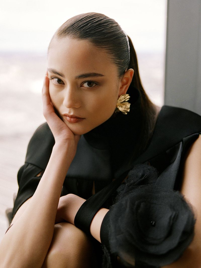 Close-up of a woman with dark hair and gold flower earrings wearing a black top, posing in front of a bright blurred background.