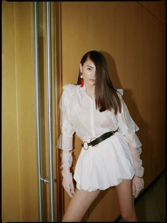 A brunette woman in a white blouse and matching skirt poses in an indoor setting with a yellow paneled wall.