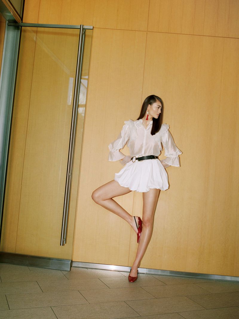 Elegant woman in a white dress and red shoes poses indoors with a light yellow wall and glass door, showcasing a fashion-forward look.