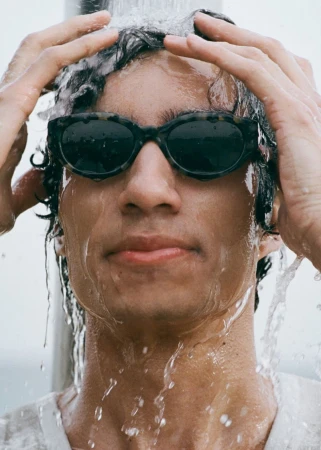 Close-up portrait of a man wearing dark sunglasses, running wet hands through his dark hair beneath cascading water.