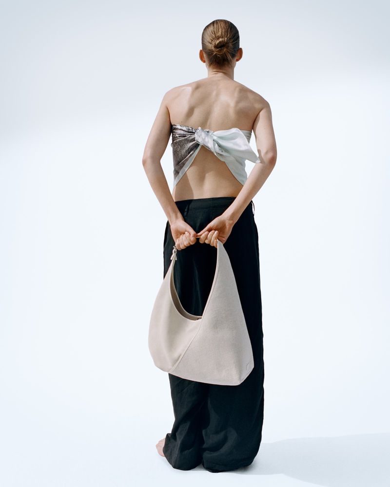Model from behind wearing a patterned backless top, dark trousers and holding a light-coloured shoulder bag on white background