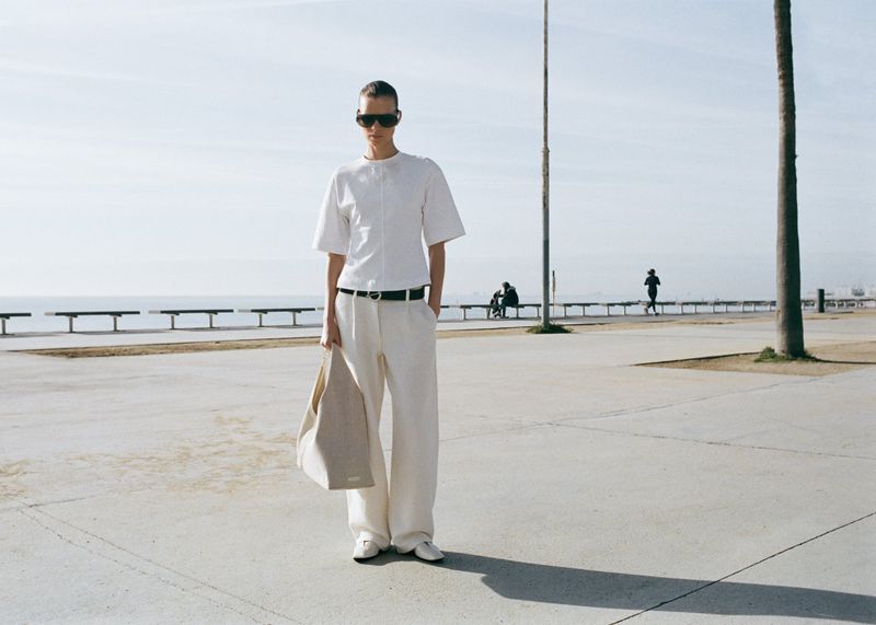 Model in white shirt and trousers wearing dark sunglasses holding a tote bag on a paved promenade by the sea