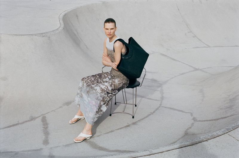 Model seated on a metal chair holding a dark tote bag inside a concrete skate bowl for Samsoe Samsoe Pre Autumn 26.