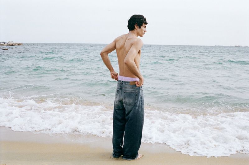 Shirtless male model wearing baggy denim jeans standing on a sandy beach looking toward the ocean