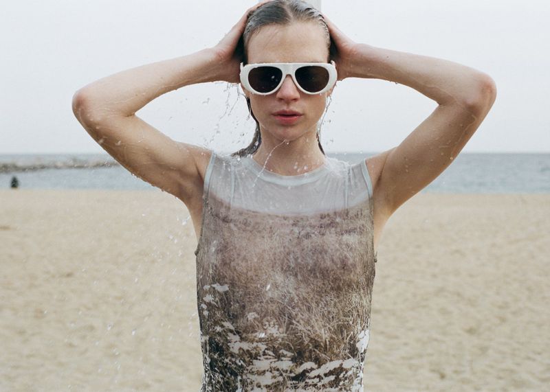 Model wearing white sunglasses and a printed sleeveless top with wet hair on a sandy beach
