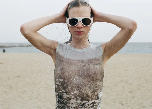 Model wearing white sunglasses and a printed sleeveless top with wet hair on a sandy beach