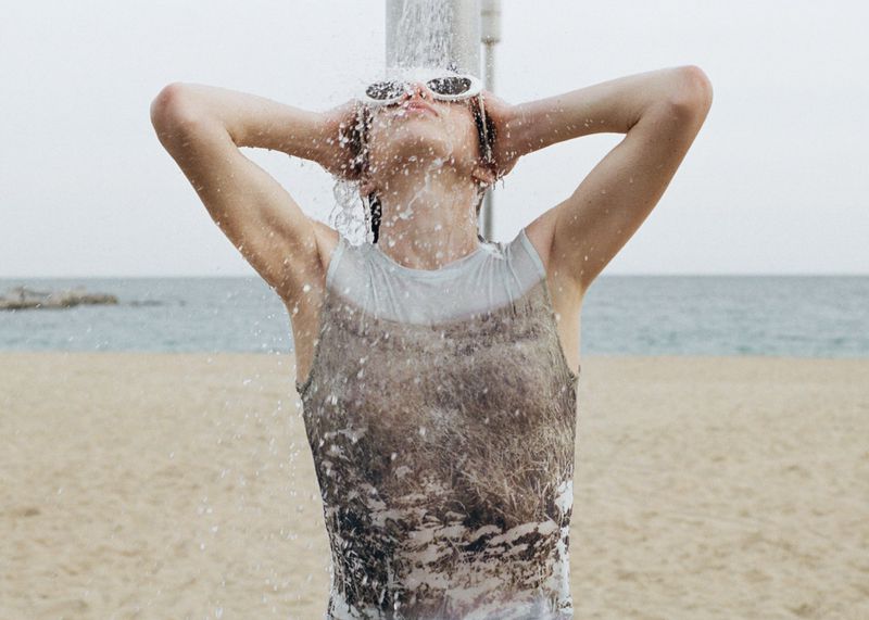 Model wearing sunglasses standing under an outdoor shower on a sandy beach with the ocean in the background
