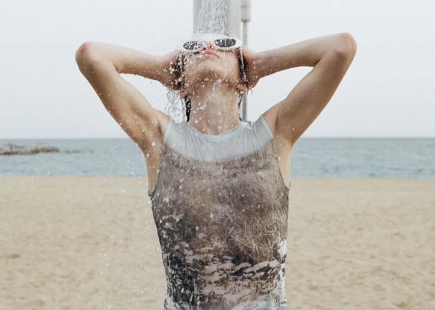 Model wearing sunglasses standing under an outdoor shower on a sandy beach with the ocean in the background