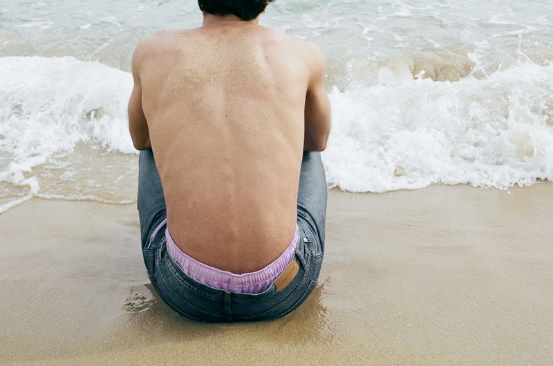 Shirtless man seated on a sandy beach with ocean waves breaking behind him, wearing denim jeans.